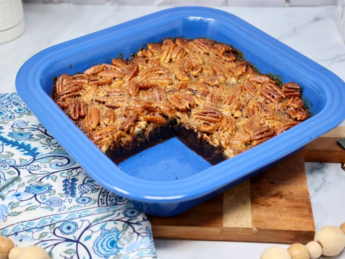 sliced pecan pie brownies in a blue baking dish on top of a wood cutting board with a blue and white patterned napkin