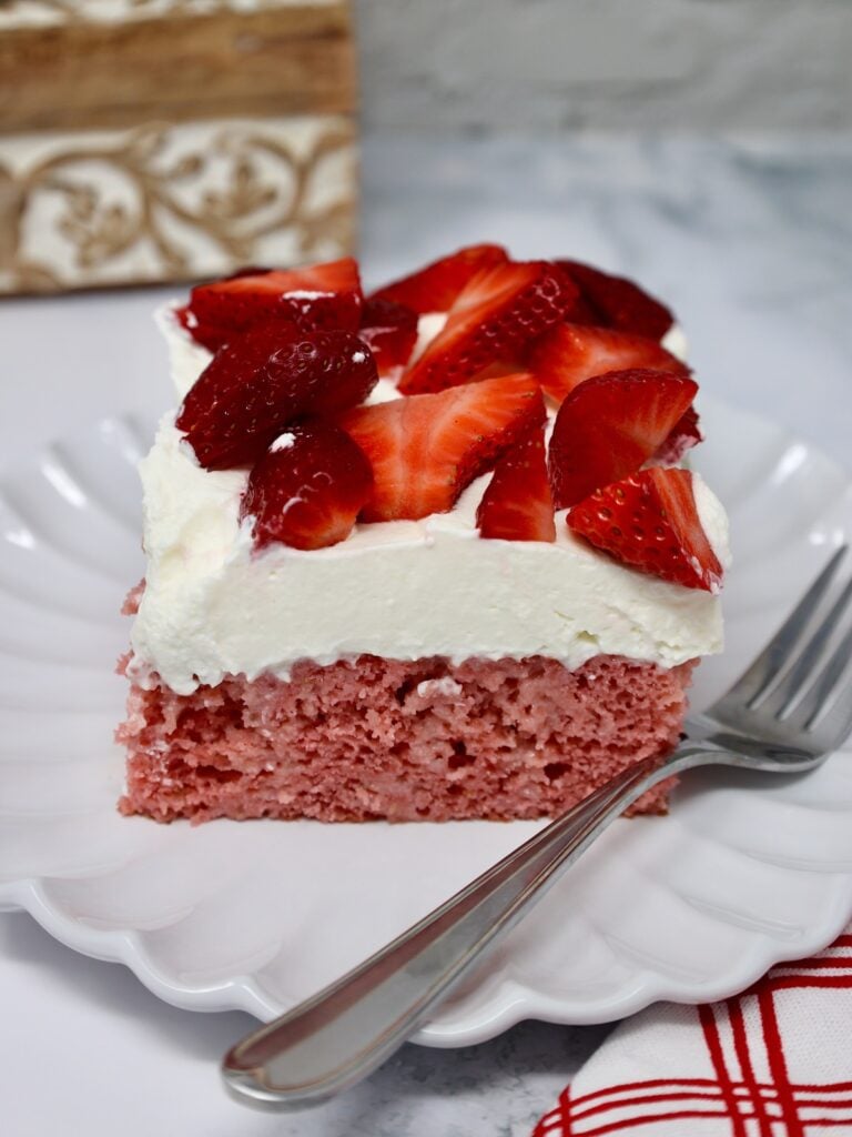 strawberries and cream sit atop a beautiful strawberry poke cake on a pretty white plate
