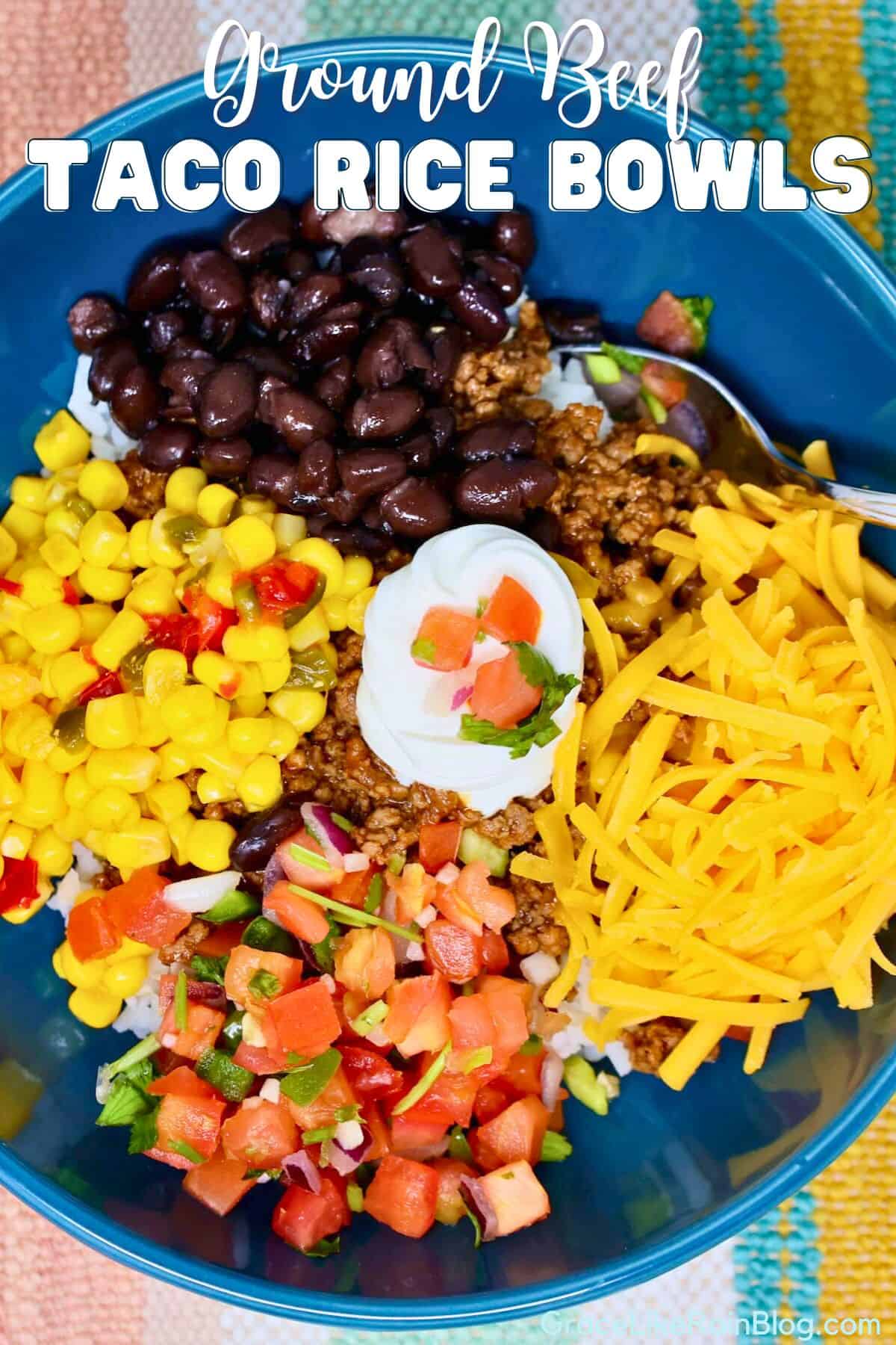 Ground beef taco rice bowls topped with cheese, lettuce, tomatoes, corn, black beans, cheese, and sour cream