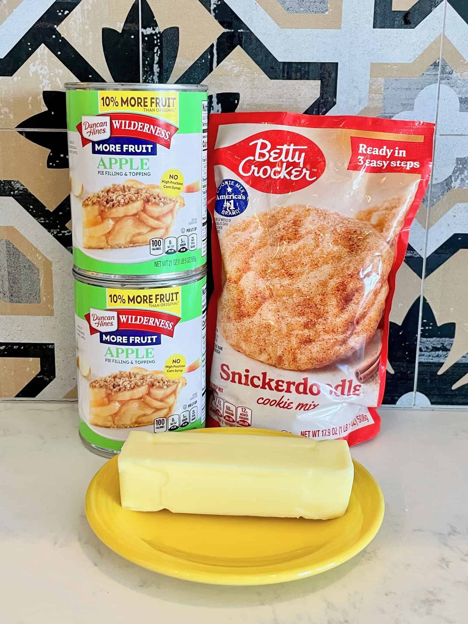 ingredients for snickerdoodle apple crisp, including apple pie filling, a package of snickerdoodle cookie mix, and butter