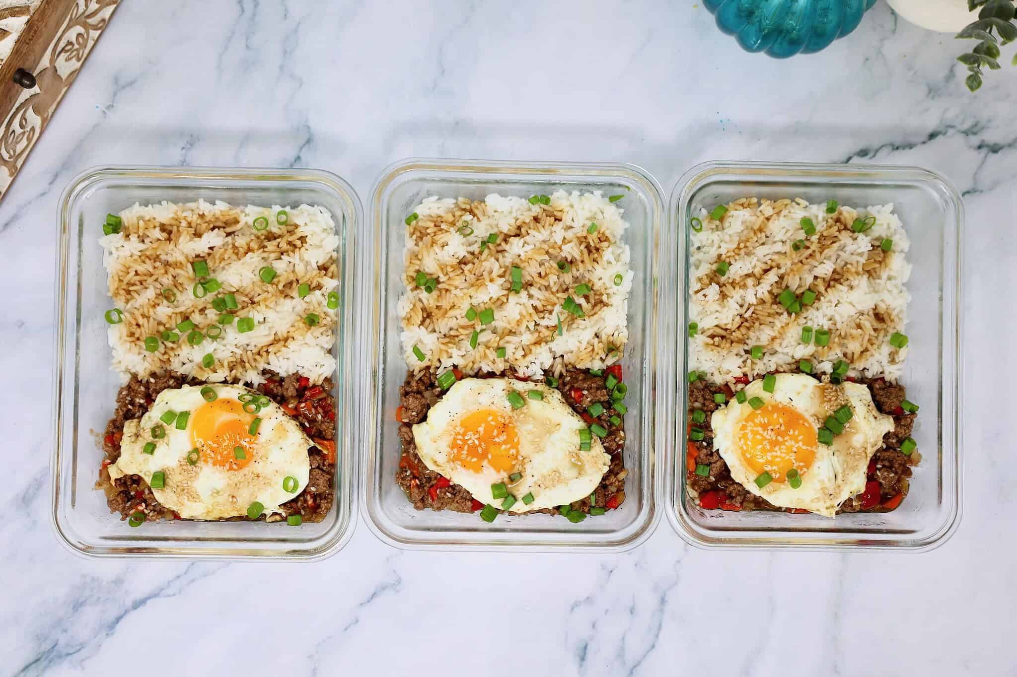 Over-medium egg being added to a teriyaki ground beef rice bowl meal prep container